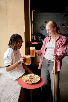 Vertical shot of young adult women drinking beer and sharing snacks beside pool table in bar, celebrating friendship and leisure. Ideal for nightlife marketing, social lifestyle campaigns
