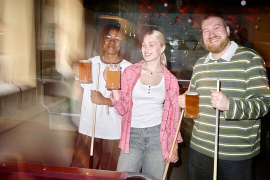 Portrait of diverse young adults holding beer mugs and pool cues, celebrating social nightlife and friendship after game. Useful for bar marketing, leisure campaigns, group bonding themes
