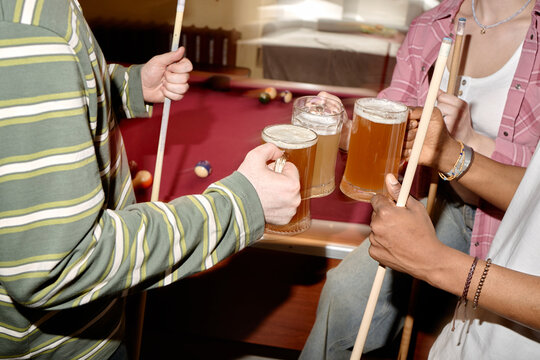 Young adults holding pool cues and clinking beer mugs beside billiards table, socializing during casual nightlife gathering. Useful for friendship, leisure, bar entertainment marketing
