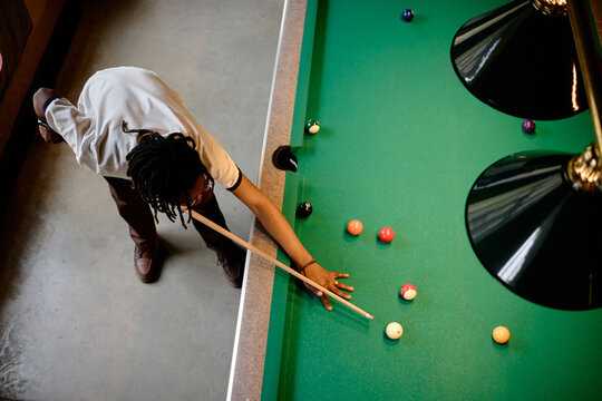 High angle shot of young Black man playing billiards, aiming cue stick over pool table during leisure activity. Suitable for lifestyle marketing, recreation venues, social campaigns

