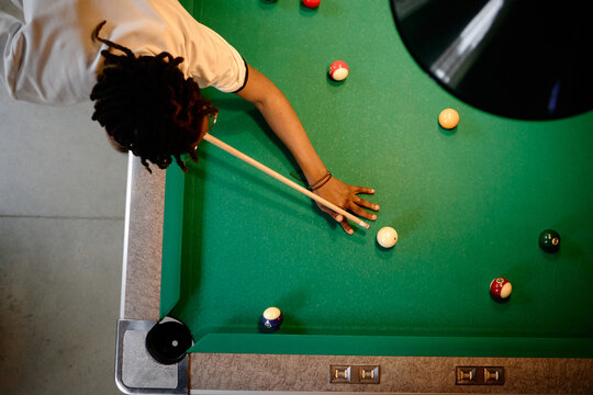 Overhead shot of young adult man playing billiards, aiming cue stick across pool table during leisure game. Suitable for recreation marketing, bar promotion, social lifestyle content
