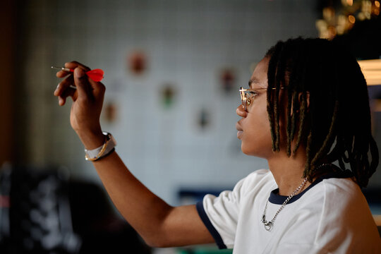 Black teenager holding dart and aiming at target, practicing focus and precision during game activity. Useful for youth concentration, leisure marketing, skill development campaigns
