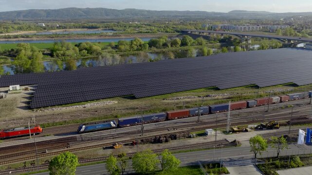 Aerial view of a large solar farm next to railway tracks with driving trains  and a highway bridge, symbolizing energy transition and modern infrastructure.