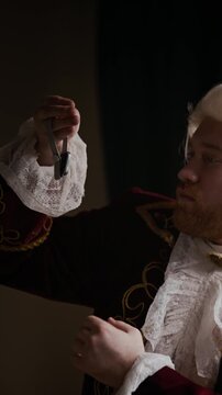 Vertical slowmo shot of Caucasian bearded man wearing baroque embroidered coat, wig and lace cravat examining smartwatch indoors