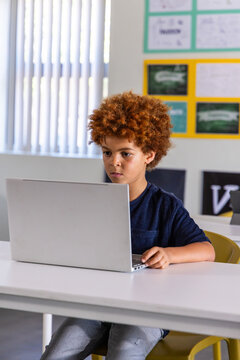 African American child boy sitting on yellow chair at white desk in classroom using silver laptop