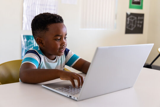 African American boy wearing striped shirt sitting in yellow chair at table typing on silver laptop