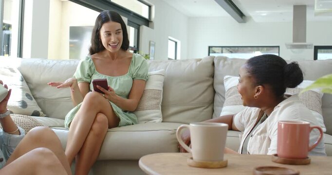 Diverse female pals on sofa, center woman showing phone and sharing post, others laughing with mugs