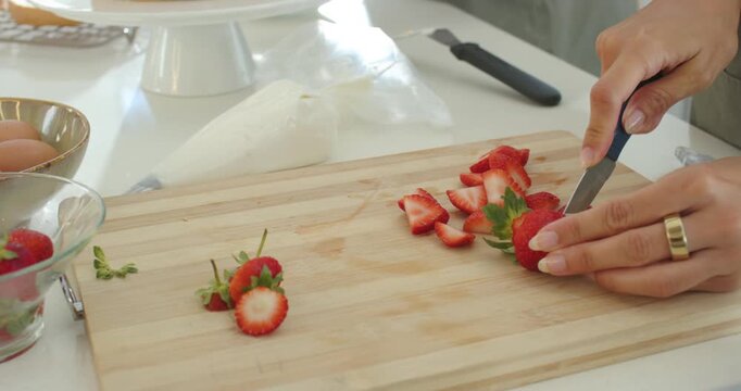 Adult women taking strawberries, slicing on board in kitchen while frosting cake to top, copy space