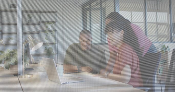 Working three coworkers leaning over open laptop at office desk, wearing pink, gray, maroon tops