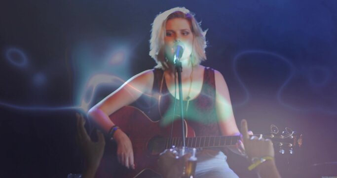 Singing teen playing acoustic guitar, mic on club stage, wearing dark tank top and light pants
