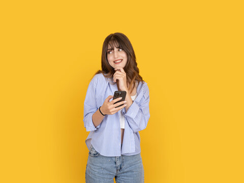 Playful young woman holding smartphone and touching chin with flirty curious smile, looking aside while considering message. Isolated on yellow background with copy space for advertising use.