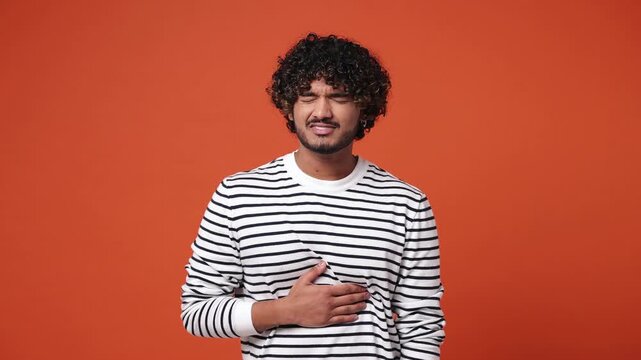 Sick ill sad young Indian man he wear striped sweatshirt casual clothes put hands on abdomen suffering from stomach-ache griping bellyache feel bad seedy isolated on plain orange red color background