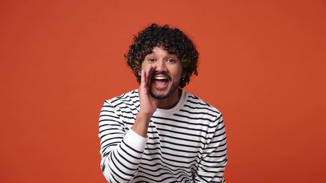 Excited happy fun young Indian man wear striped sweatshirt casual clothes scream hot news about sales discount with hands near mouth hurry up isolated on plain orange red background. Lifestyle concept