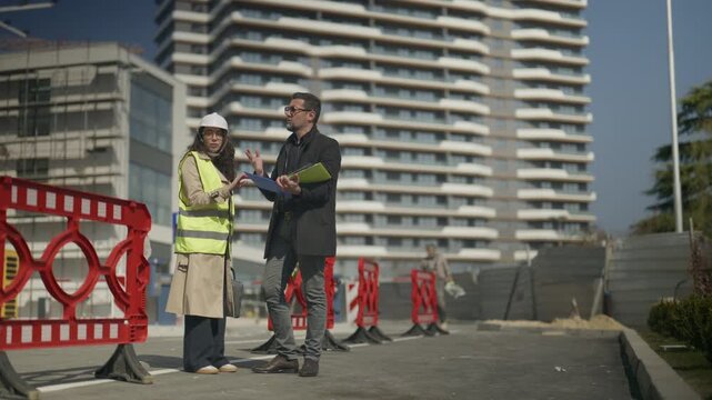Two professional architects discuss building plans while standing at a modern construction site. They review documents and collaborate on the development of a new urban project.