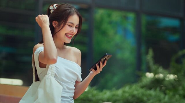 Excited young Asian woman celebrating success while looking at her smartphone in a modern city park. Concept of winning, receiving good news, stock trading profits, or viral social media success. Vibr