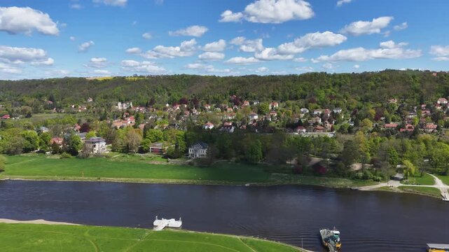 Aerial panorama of the Elbe river in Pirna featuring green riverbanks, villas on a forested hillside, and a white pier under a blue cloudy sky.