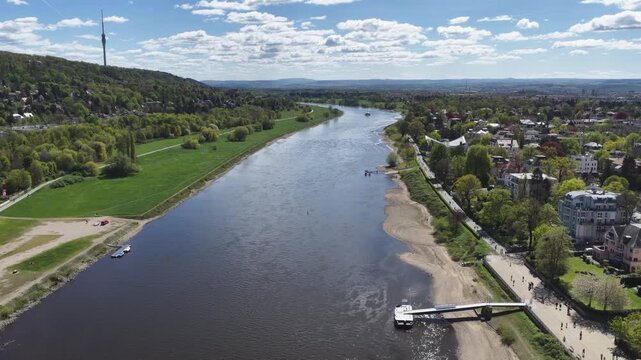 Aerial view of the Elbe river near DresdenBlasewitz  featuring the television tower, a steamboat pier, and green riverbanks under a blue cloudy sky in spring.
