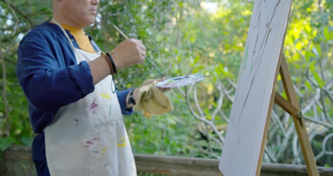 Senior Asian painter wearing stained apron mixing on palette, painting composition on easel on deck
