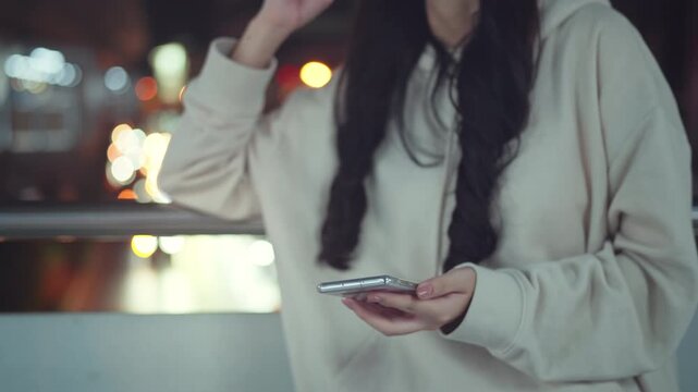 Closeup - Happy young Asian woman in a cozy hoodie using her smartphone on a city bridge at night. Beautiful urban bokeh lights in the background. Concept of modern connectivity, social media usage, a