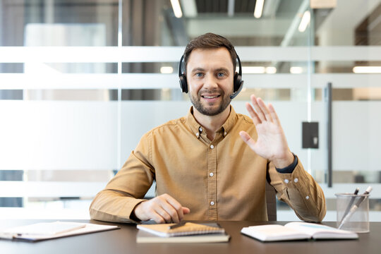 Man wearing a headset and brown shirt smiling, waving hello to the camera, and engaging in online communication with clients or colleagues while sitting at an office desk