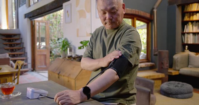Senior Asian man in olive t-shirt prompted by phone adjusting BP cuff with monitor in kitchen