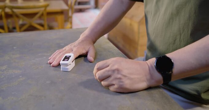 Senior man noticing lit oximeter, reaching, placing finger and measuring oxygen on kitchen counter