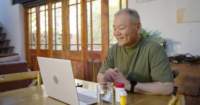 Senior Asian man joining call, laughing, inspecting pill bottle and showing label to laptop at home