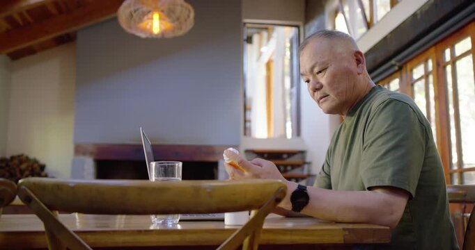 Senior Asian man in green shirt sitting at home comparing prescriptions using laptop, copy space