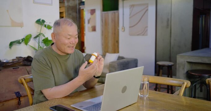 Senior Asian male at home holding orange bottle, presenting white bottle red lid for laptop consult