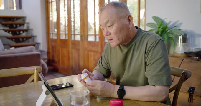 Senior Asian man sitting at dining table, holding pill bottle, showing label to tablet call