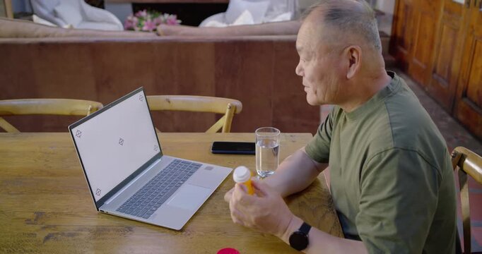 Senior Asian man sitting at wooden table reading Rx label following laptop prompts, copy space
