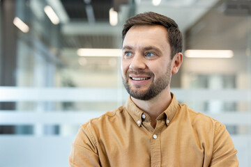 Naklejka premium Happy bearded professional man in a casual yellow shirt smiling and looking away confidently in a bright modern office interior, embodying optimism and career success