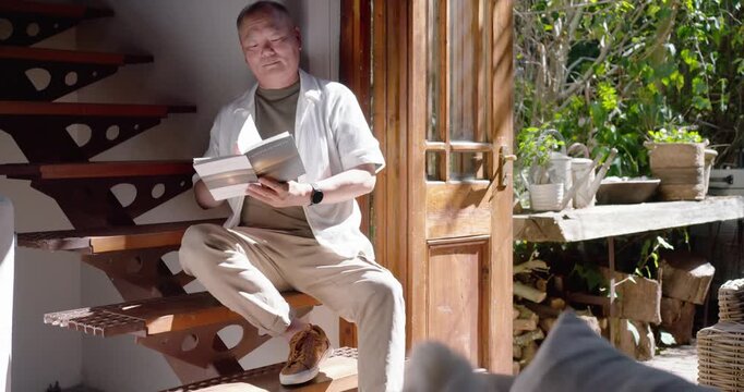 Senior Asian man sitting on spiral step, sun drawing gaze, pausing reading book, glancing at garden