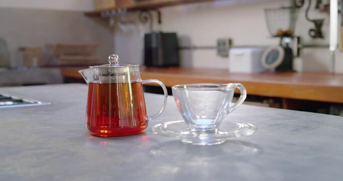 Camera is pulling back and tilting up, revealing glass teapot and empty teacup on grey counter