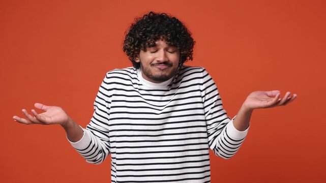 Hesitant indecisive fun shy sad young Indian man he wears striped sweatshirt casual clothes feels doubtful spreading hands say oops ouch oh omg i am so sorry isolated on plain orange red background
