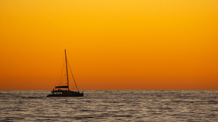 Obraz premium Silhouette of a distant Catamaran on the ocean against a vibrant golden orange sunset off the coast at Cape Town South Africa.