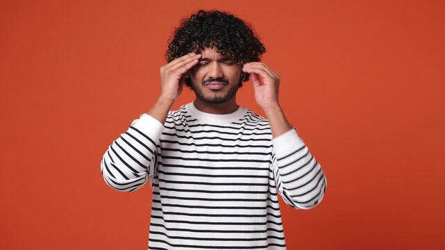 Sick ill sad tired young Indian man wear striped sweatshirt casual clothes put hands on head rub temples having headache suffering from migraine feel bad seedy isolated on plain orange red background