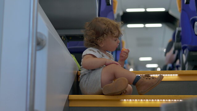 Toddler with curly hair sitting on train steps eating snack with blue seats and bright interior lights in background during journey