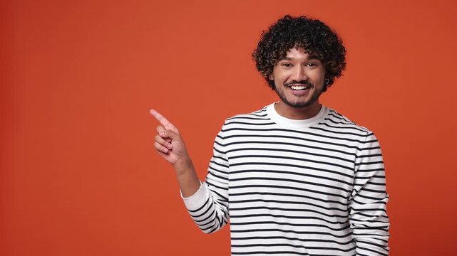 Young Indian man he wear striped sweatshirt casual clothes pointing indicate index finger aside on workspace area copy space isolated on plain orange red background studio portrait. Lifestyle concept