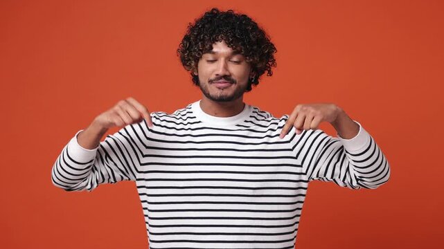 Young Indian man he wear striped sweatshirt casual clothes pointing indicate index finger down on workspace area copy space isolated on plain orange red background studio portrait. Lifestyle concept