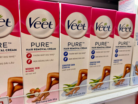 Rows of Veet Pure hair removal cream boxes are displayed on a store shelf. The packaging features a pink and white color scheme. Each box highlights different skin types and purposes.