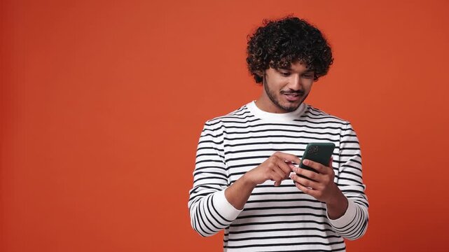 Young Indian man he wear striped sweatshirt casual clothes hold in hand use mobile cell phone indicate index finger aside on workspace area copy space mock up isolated on plain orange red background