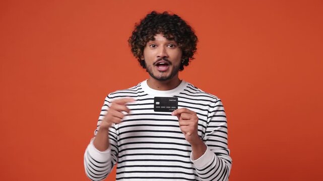 Young Indian man wear striped sweatshirt casual clothes hold in hand pointing index finger on mockup plastic credit bank card show thumb up isolated on plain orange red background. Lifestyle concept