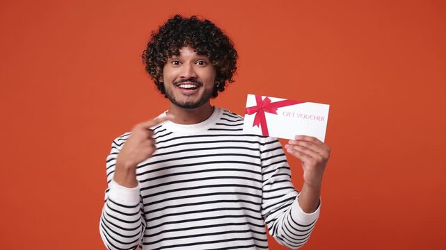 Young Indian man wear striped sweatshirt casual clothes hold in hand pointing index finger on mockup plastic credit bank card showing thumb up isolated on plain orange red background studio portrait
