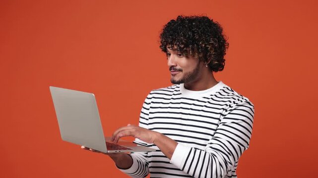 Young smart IT user Indian man he wear striped sweatshirt casual clothes hold in hand use work typing on laptop pc computer isolated on plain orange red background studio portrait. Lifestyle concept