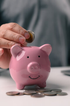 Adult hand inserts silver coin inside slot pink ceramic piggy bank sitting white table surrounded scattered change indoor setting representing financial planning retirement security investment.