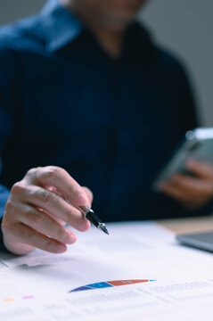 Businessman holds pen over financial report pie chart gripping smartphone office setting evening work session analyzing corporate data statistics professional finance management executive strategy.