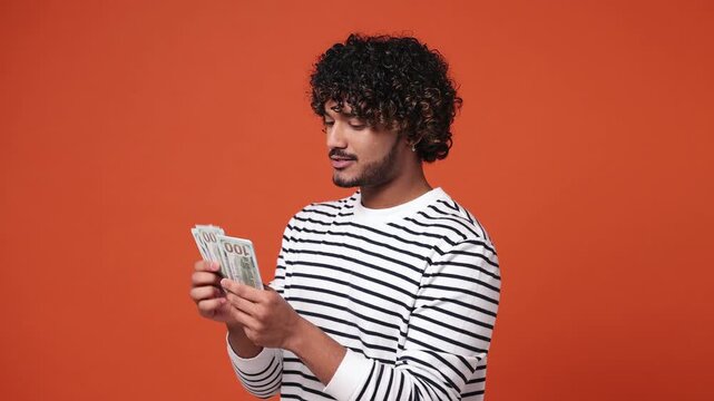 Happy accountant young Indian man he wear striped sweatshirt casual clothes hold in hand count stack of cash money in dollar banknotes isolated on plain orange red background studio. Lifestyle concept