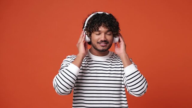 Young happy Indian man wear sweatshirt casual clothes listen mp3 music in headphones slow dance sing song have fun enjoy relax show thumb up isolated on plain orange red background. Lifestyle concept