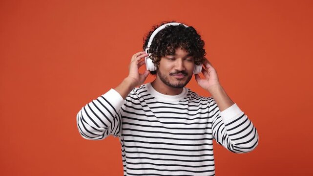 Vivid young Indian man he wearing striped casual clothes listen mp3 music in headphones dance sing song fooling around have fun enjoy relax isolated on plain orange red background. Lifestyle concept